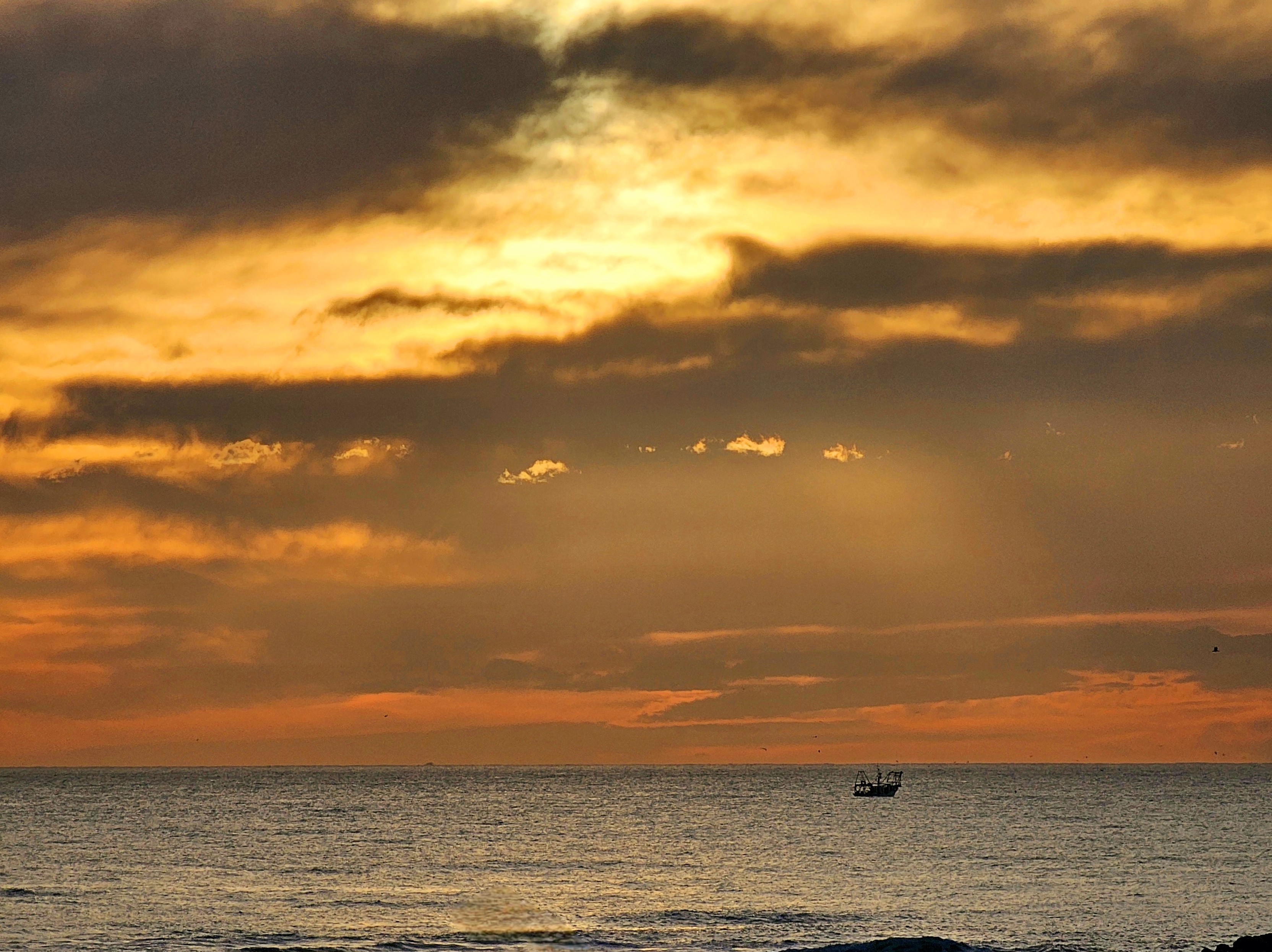 A serene sunset over the ocean, with the sky painted in warm hues of gold, amber, and soft orange. Dramatic clouds are scattered across the sky, with rays of sunlight piercing through them, casting a heavenly glow. A lone fishing boat sits on the tranquil sea, silhouetted against the vibrant colors of the dusk sky, evoking a sense of peaceful solitude.