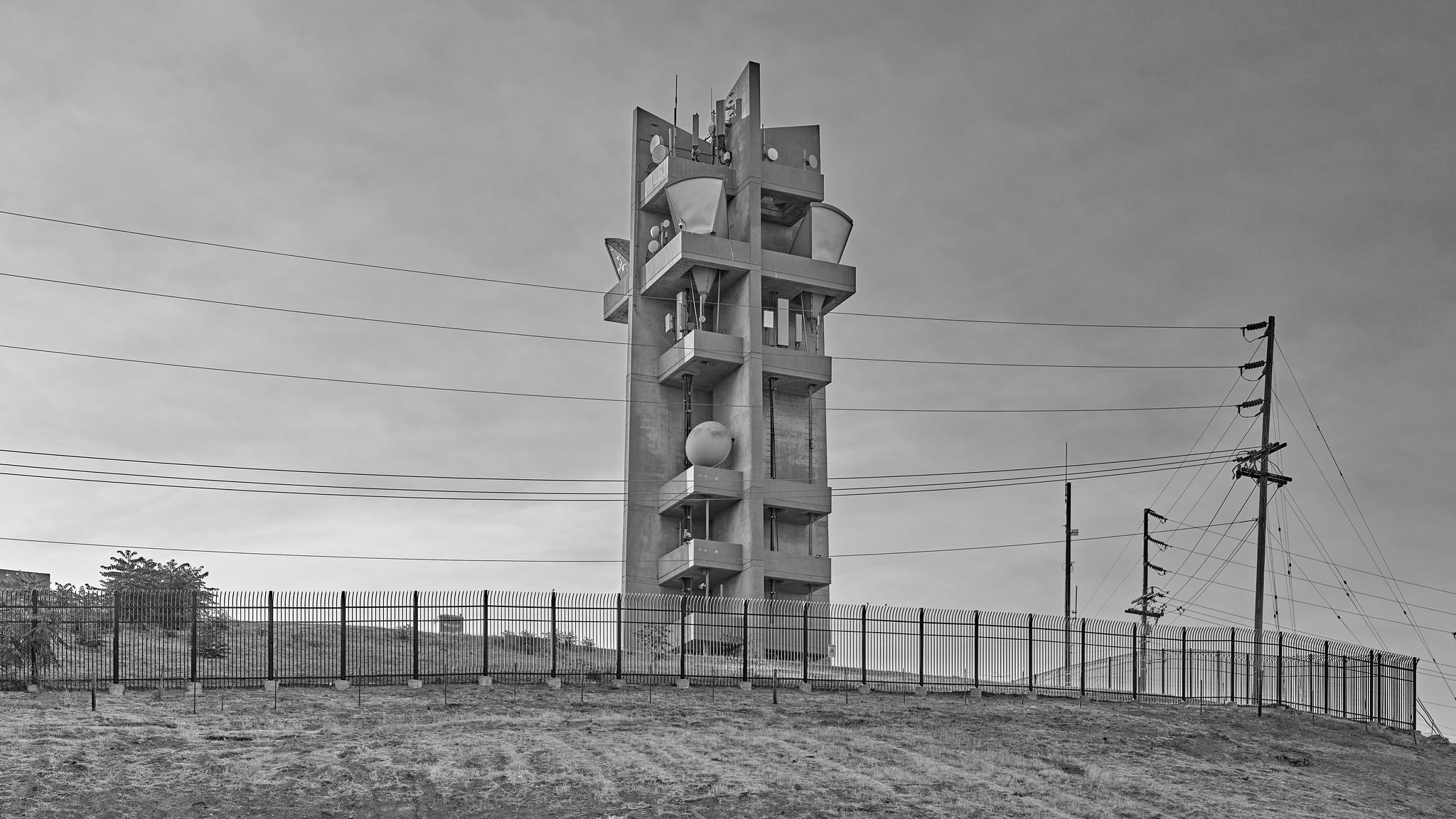 A heavy-duty Brutalist-style concrete tower, with multiple levels of platforms supporting various microwave antennas, including "horn" antennas, on a hilltop behind a fence.