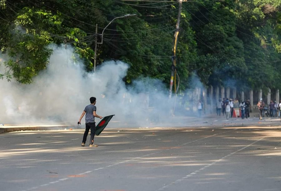 A young protester holding a Bangladesh flag stands on an empty road surrounded by thick clouds of smoke, possibly tear gas. The scene takes place in a tree-lined area with a group of people, some wearing helmets, gathered in the background. The road is hazy and tense, suggesting a moment of confrontation or unrest.