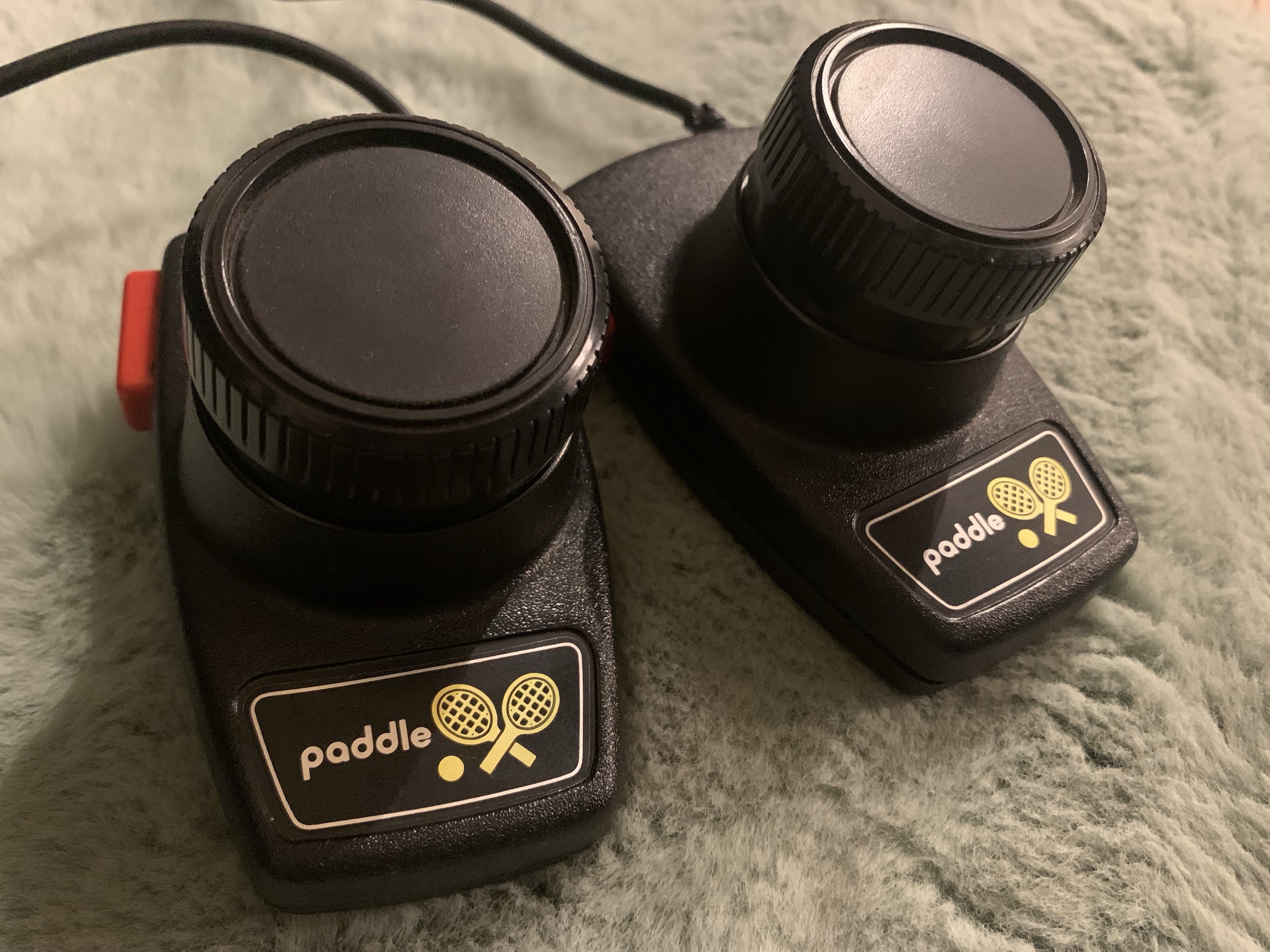 Two black paddle tennis controllers with a circular knob on top and a small red button on one side. Each features a logo with crossed paddles. The controllers are positioned on a soft, textured surface.