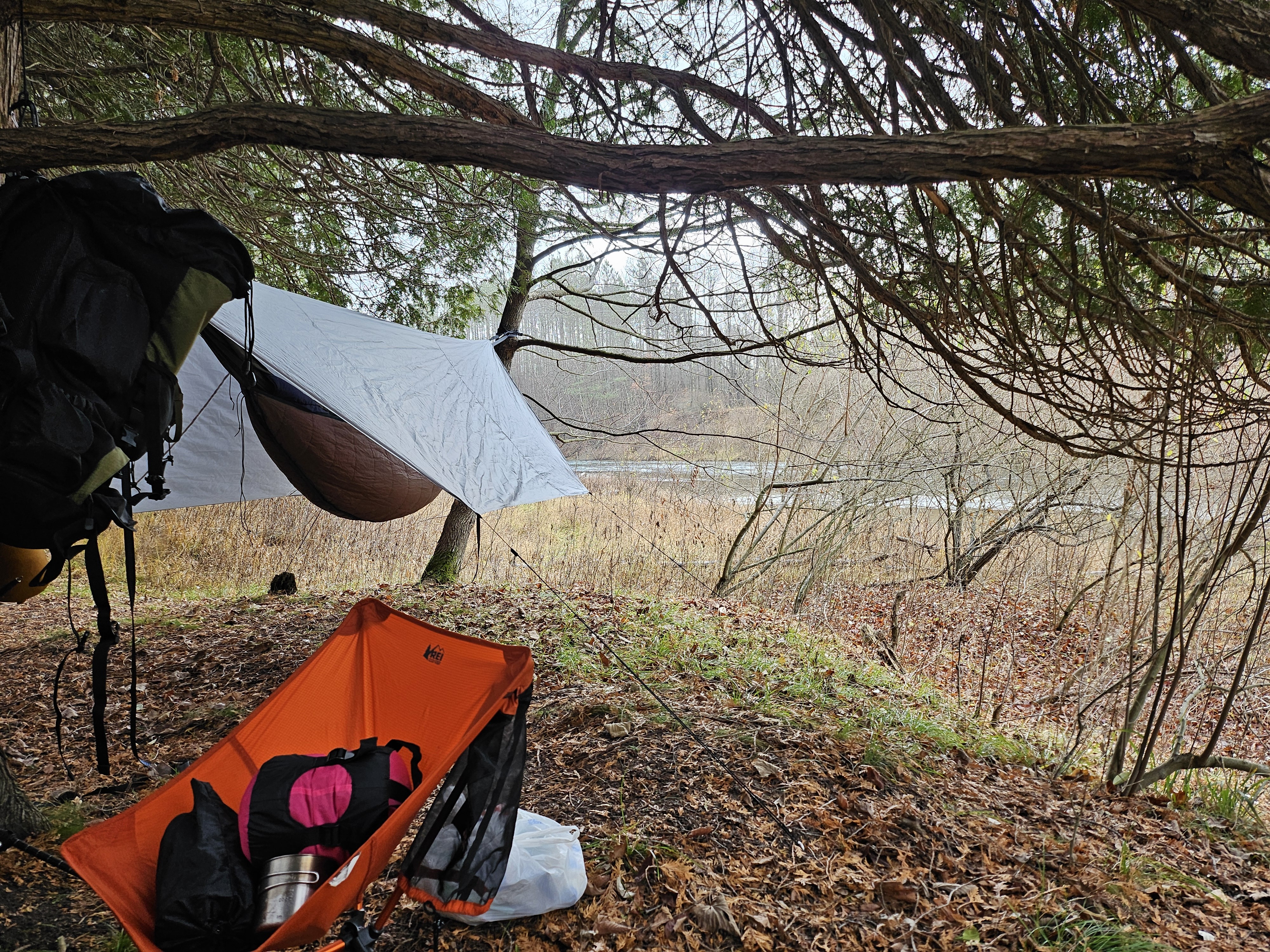 Closeup of hammock with rain fly. Orange chair on ground with items on it, backpack hanging from low tree branch 