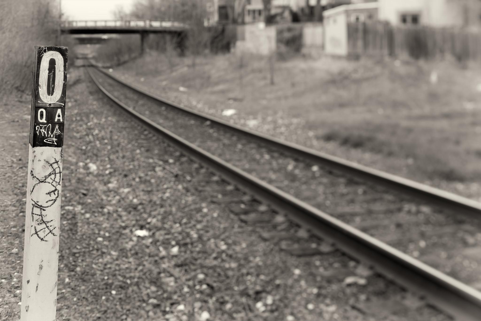 A single railroad track, receding into the distance, in a lonely urban area behind some houses. Almost everything is in soft focus. A railroad milepost, marked "O QA" is in sharp focus at left. There's a hobo-style graffiti tag on the milepost.