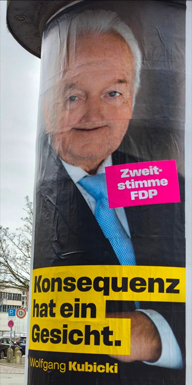 An einer Litfaßsäule hängt ein Plakat mit einem Bild von Wolfgang Kubicki und der Aufschrift "Zweitstimme FDP. Konsequenz hat ein Gesicht. Wolfgang Kubicki". Das Plakat wurde schlecht angeklebt, sodass Kubickis Gesicht auf komische Weise verzehrt ist, ähnlich wie in dem "Olol Scholol-"MichMich.