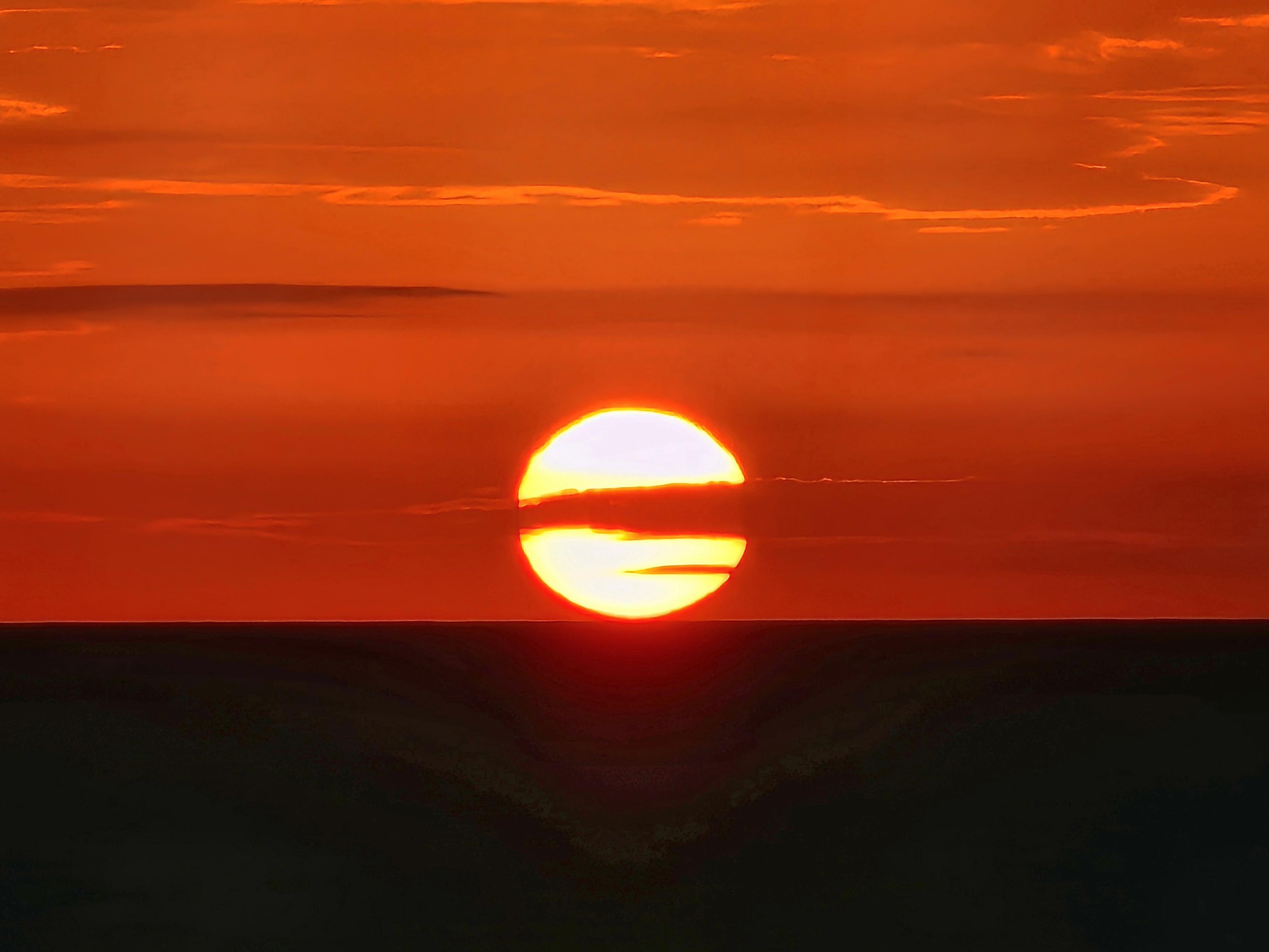 A vivid sunrise over the sea, where the sun sits low on the horizon, glowing with intense orange and red hues. The bright orb is partially obscured by thin clouds, casting warm rays across the sky. The contrast between the fiery sky and the dark ocean below creates a serene and powerful scene, symbolizing the calm transition between night and day.