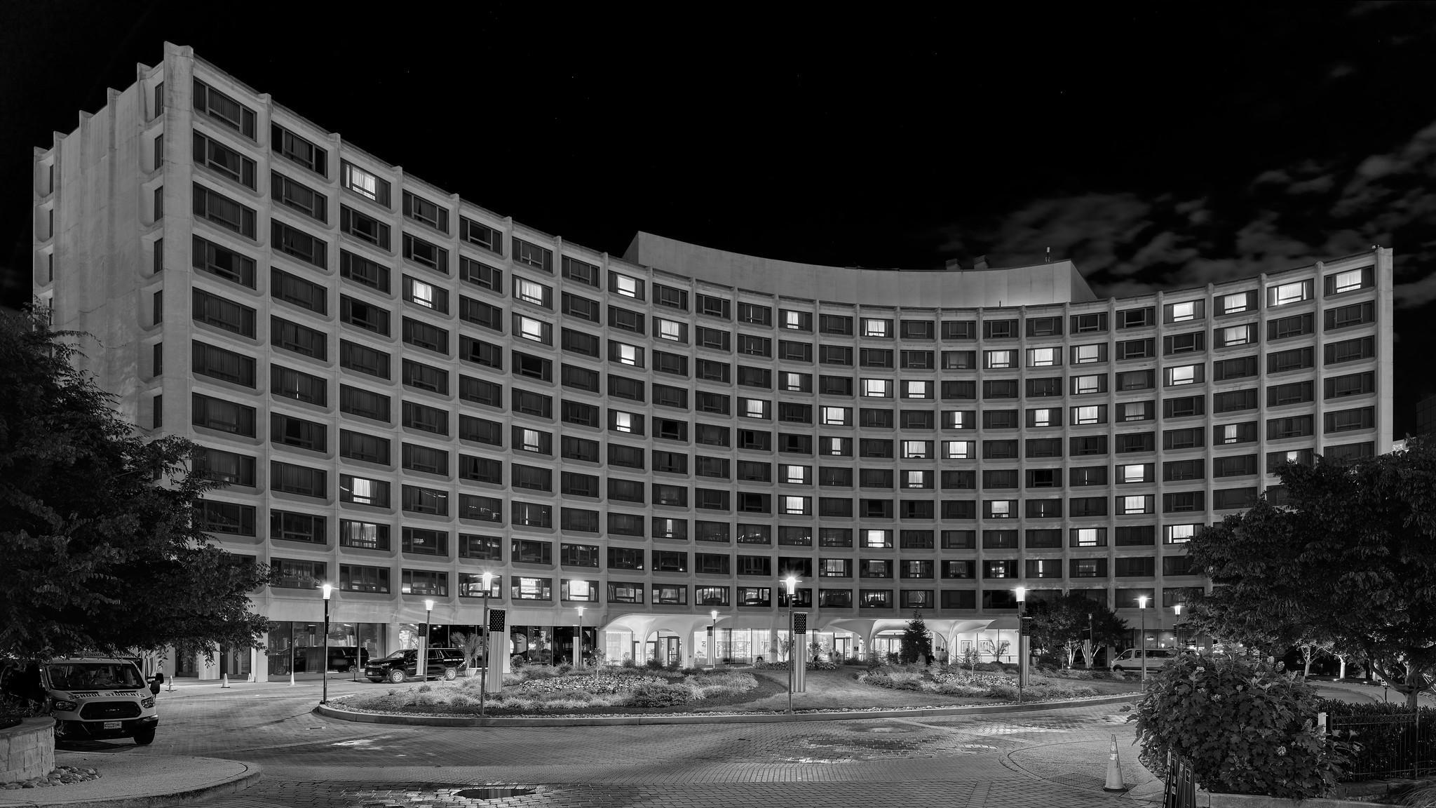 An 11 story midcentury modernist / brutalist hotel building. It has a curved facade and a plaza/driveway in front. It is night, and lights are visible in some of windows.