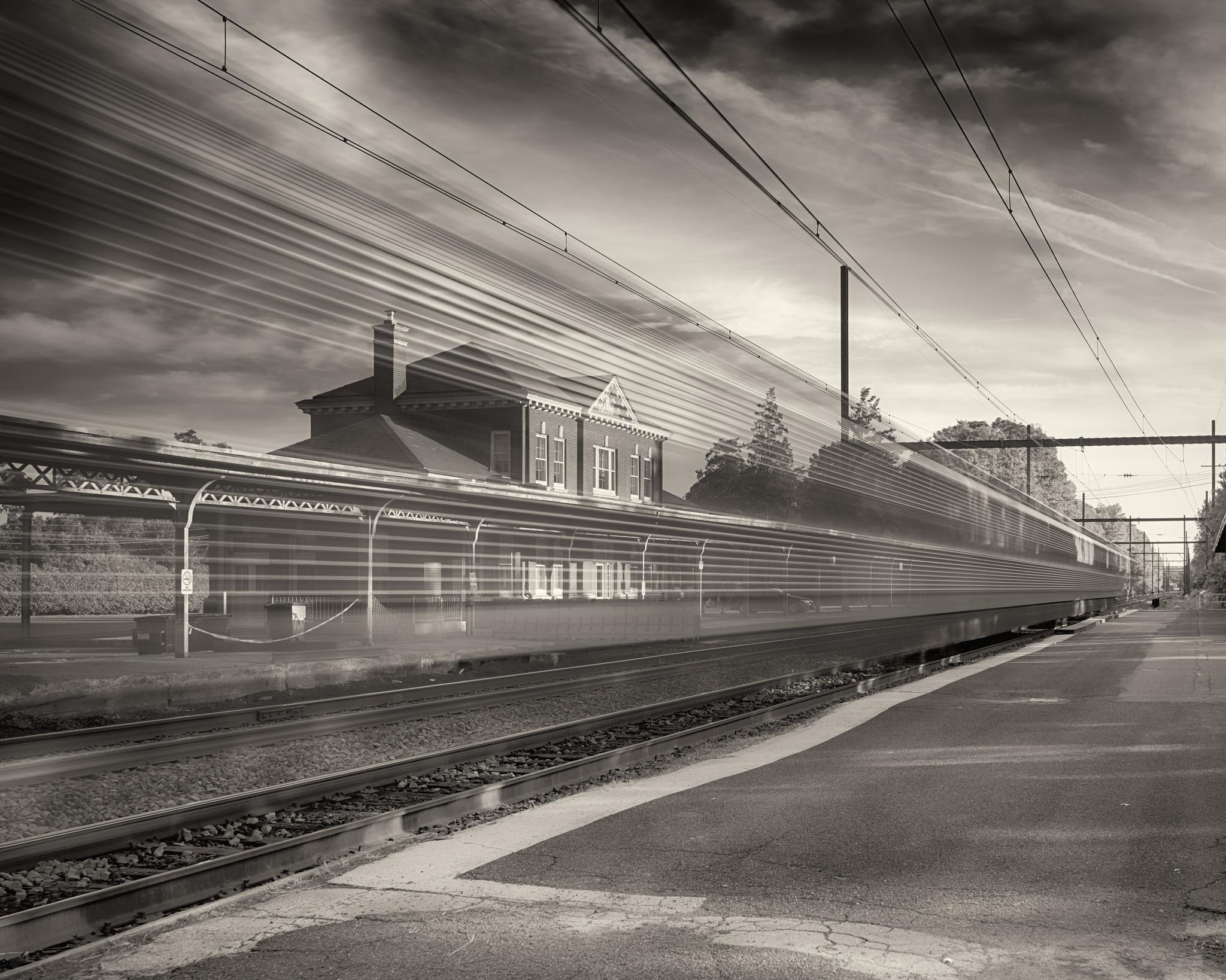 An early 20th century passenger train station, with an awning over the platform, across tracks. A blurred train streaks by in foreground.