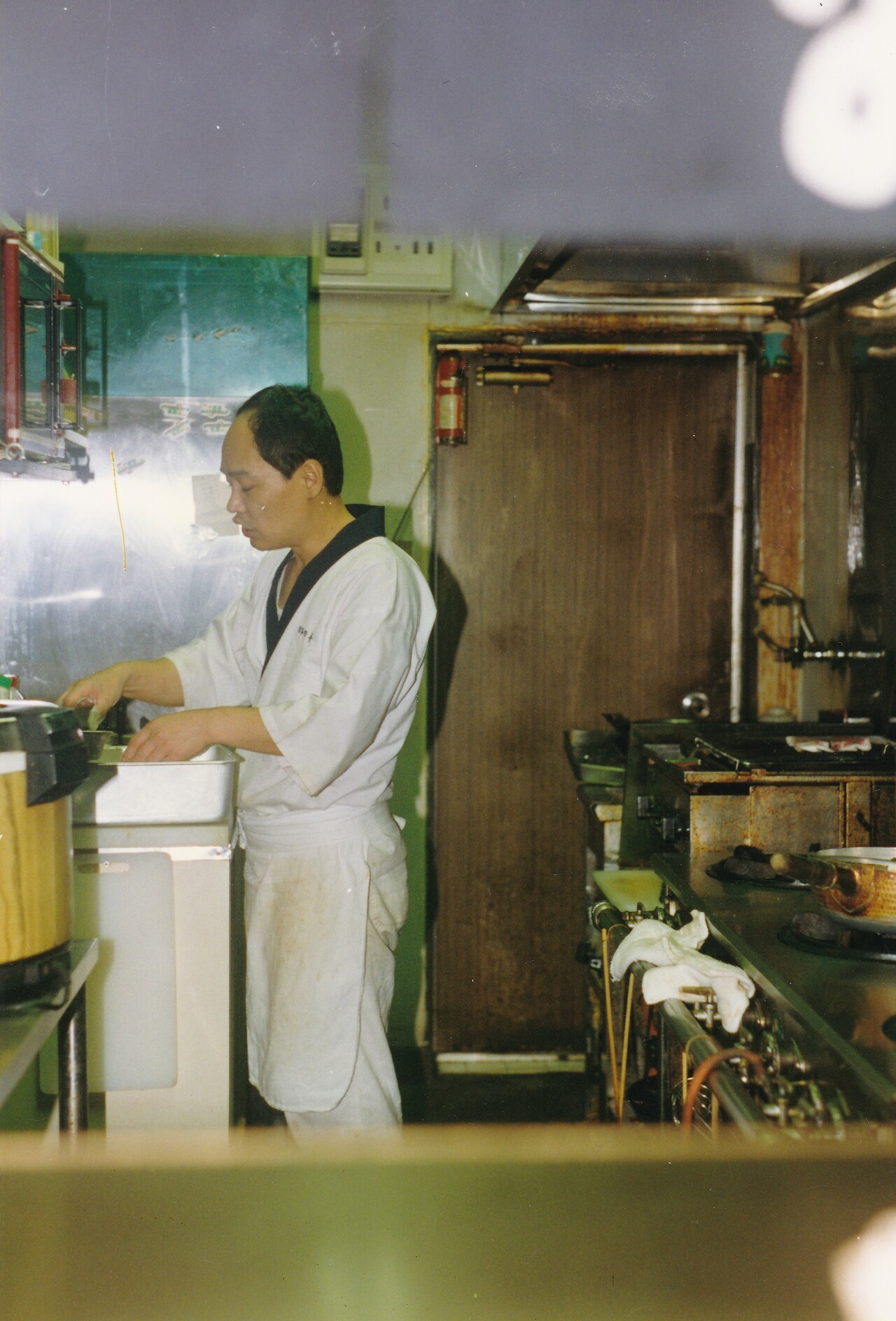 Cuisinier japonais vêtu de blanc s'affairant dans la cuisine d'un petit restaurant, photo prise en 1992