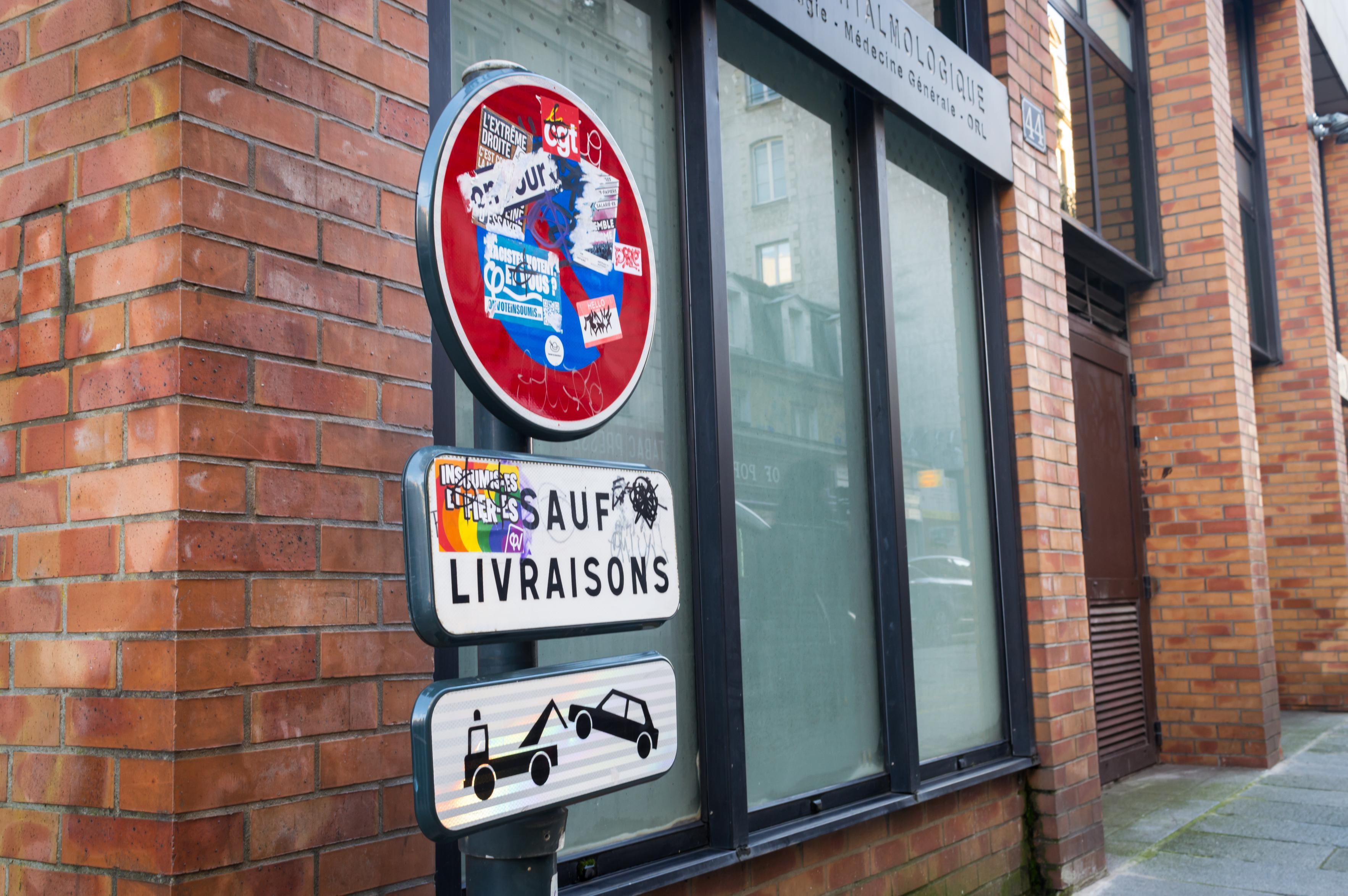 "No Parking" street sign covered in various stickers. White exception sign underneath contains a pro-LGBT sticker that has been vandalized with a black sharpie