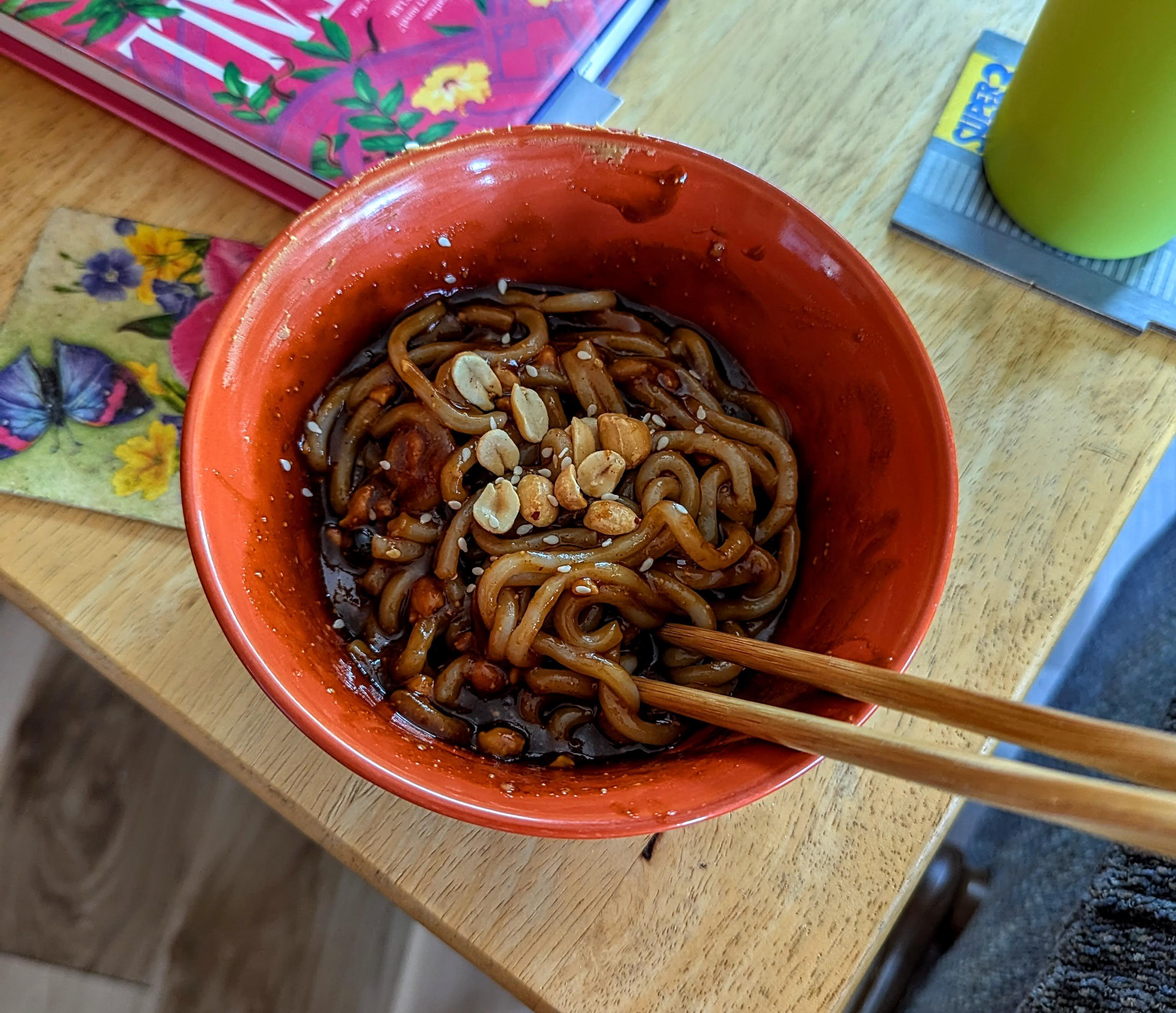 an orange bowl with a dark, sloppy sauce, from which emerge the sight of noodles topped with chopped peanuts