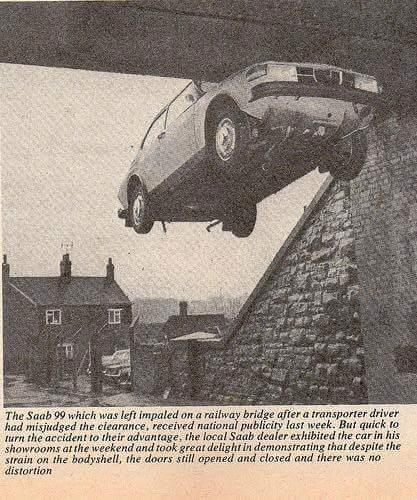 A vintage Saab 99 car is impaled on a railway bridge, seemingly suspended mid-air after an accident caused by a transporter misjudging the clearance. Despite the unusual and precarious position, the car appears intact, with its doors and bodywork showing no visible signs of damage or distortion. The photo includes a small village setting in the background, with brick houses and a cloudy sky, creating a stark contrast to the peculiar incident. A caption below the image explains that the local Saab dealer used the incident for publicity, showcasing the car in their showroom to highlight its durability.