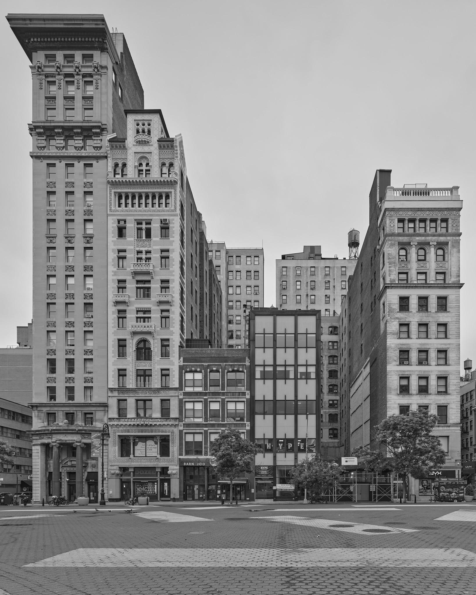 Facades of five late 19th- and early 20th- century commercial and residential buildings on a city block, with a plaza in front.