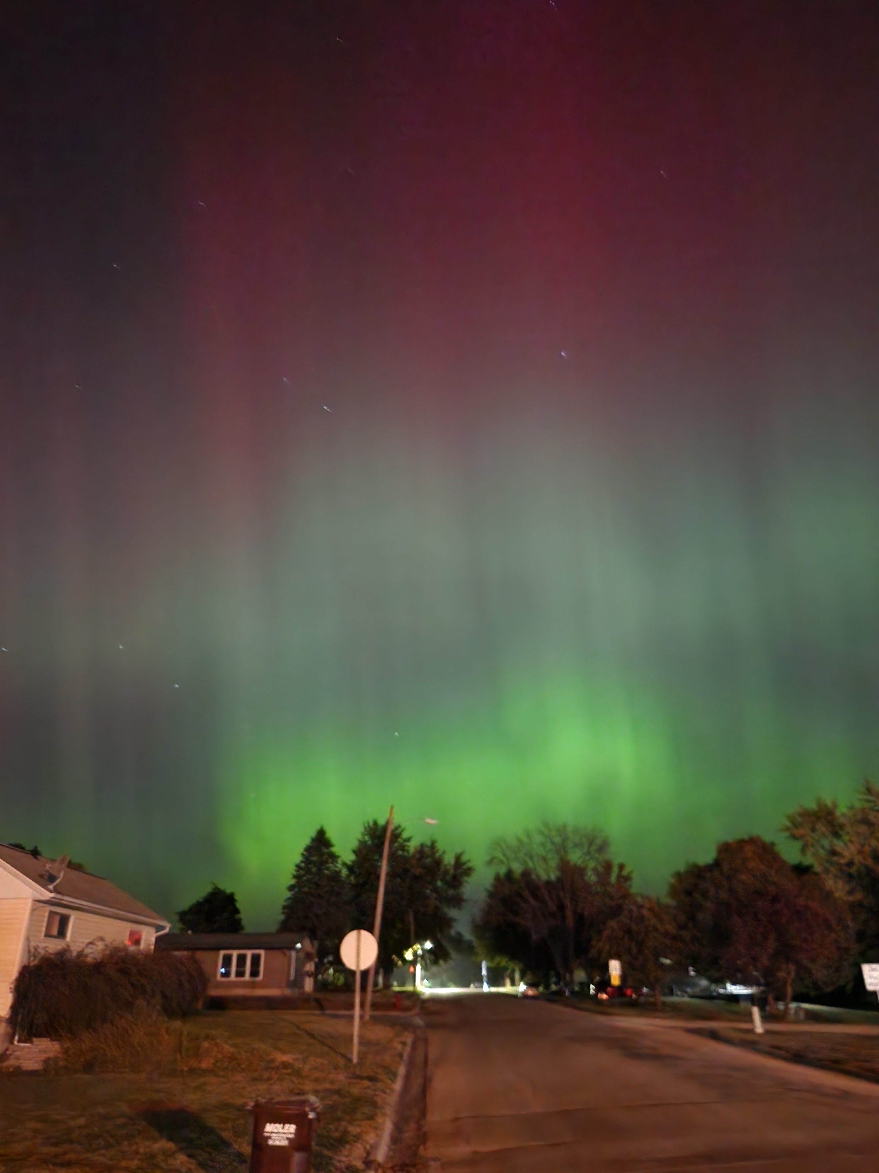 a long exposure of a street scene at night. in the sky are green, teal, and red aurora bands