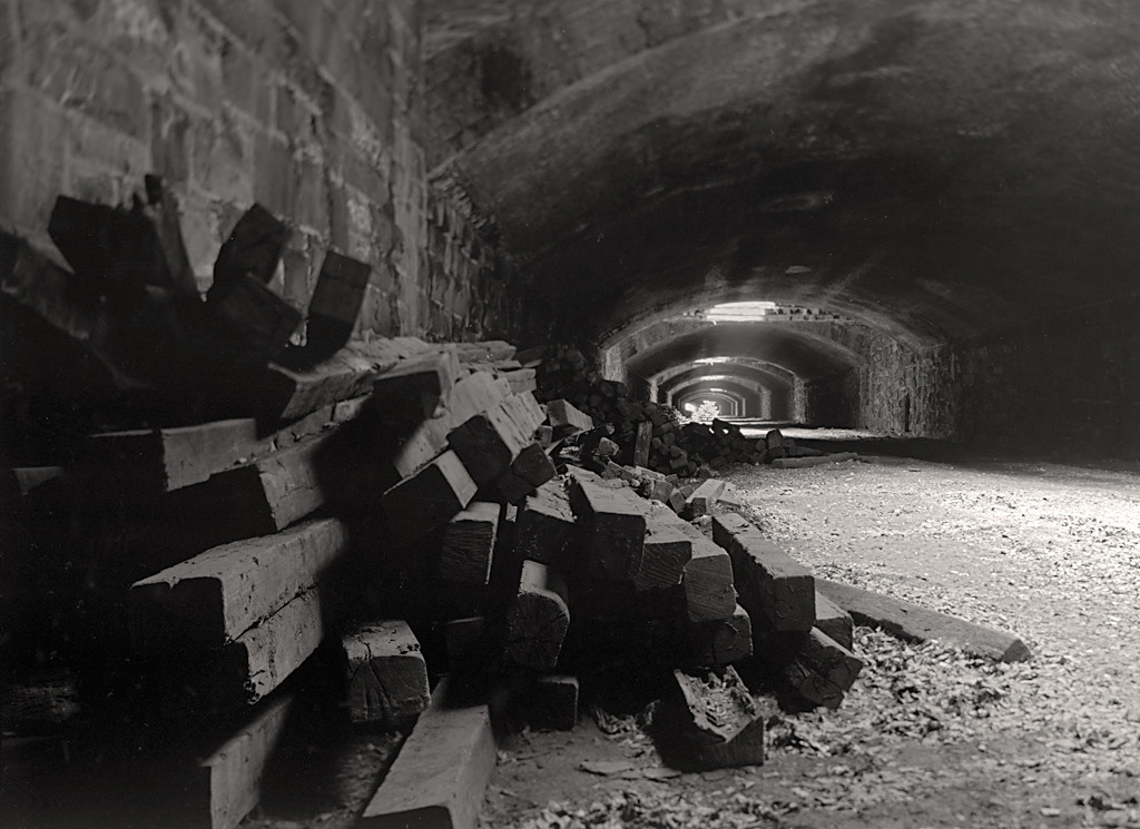 Interior of abandoned railroad tunnel with arched ceilings and grates admitting light at intervals. A stack of railroad ties is in foreground left.