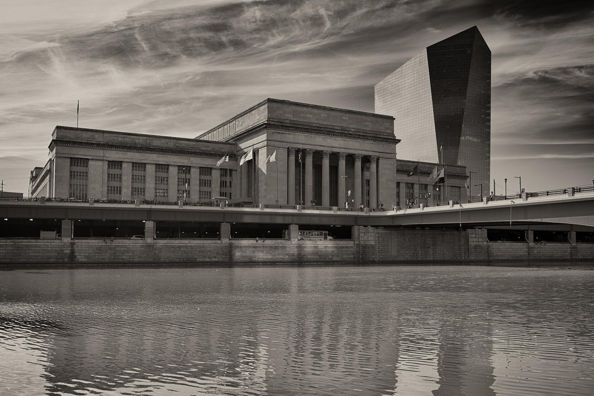 A large neoclassical train station across a river. To the right of the station is a modernist glass office tower.