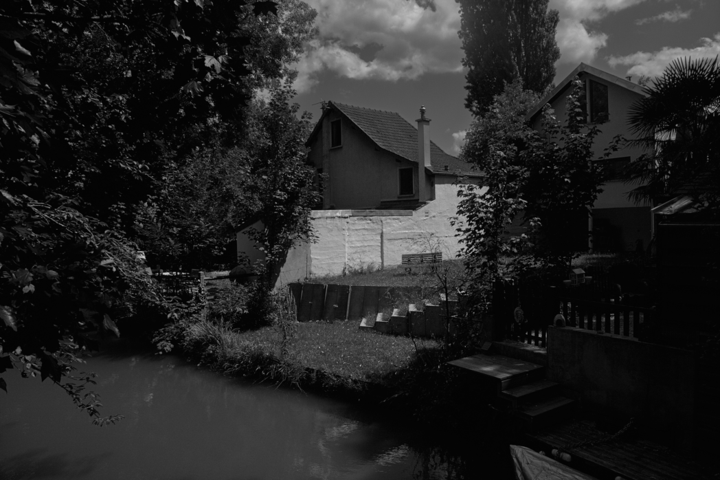 Black and white daylight outdoor photo.
Escher hidden somewhere behind the greenery curtains of one's urban mind. Of course a bench is schooling a river about the advantages of being liberated from expectancy theories.