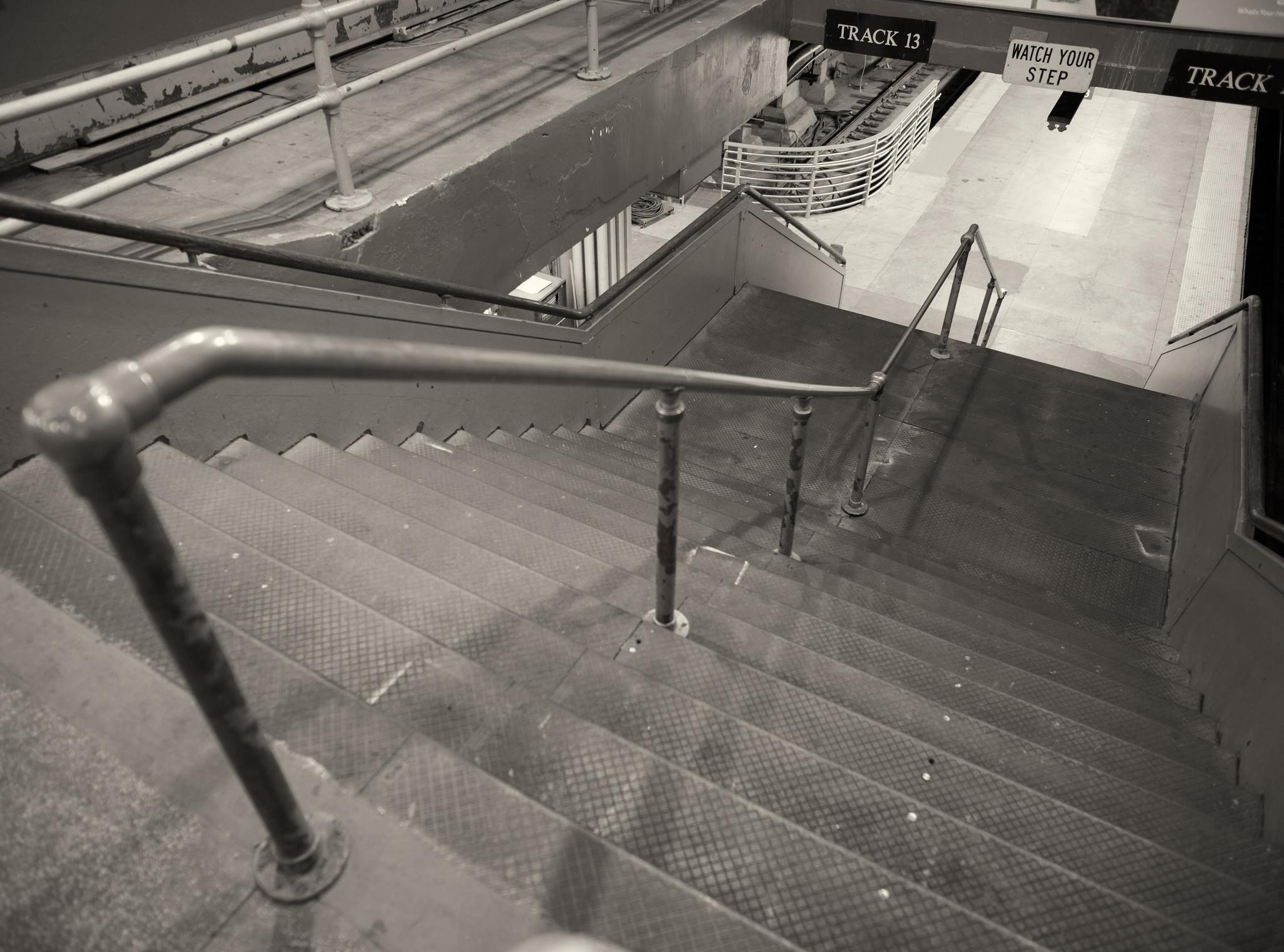 A staircase, with a simple pipe handrail at center, descending to a trail platform. Signs above read "Track 13" and "Watch Your Step".