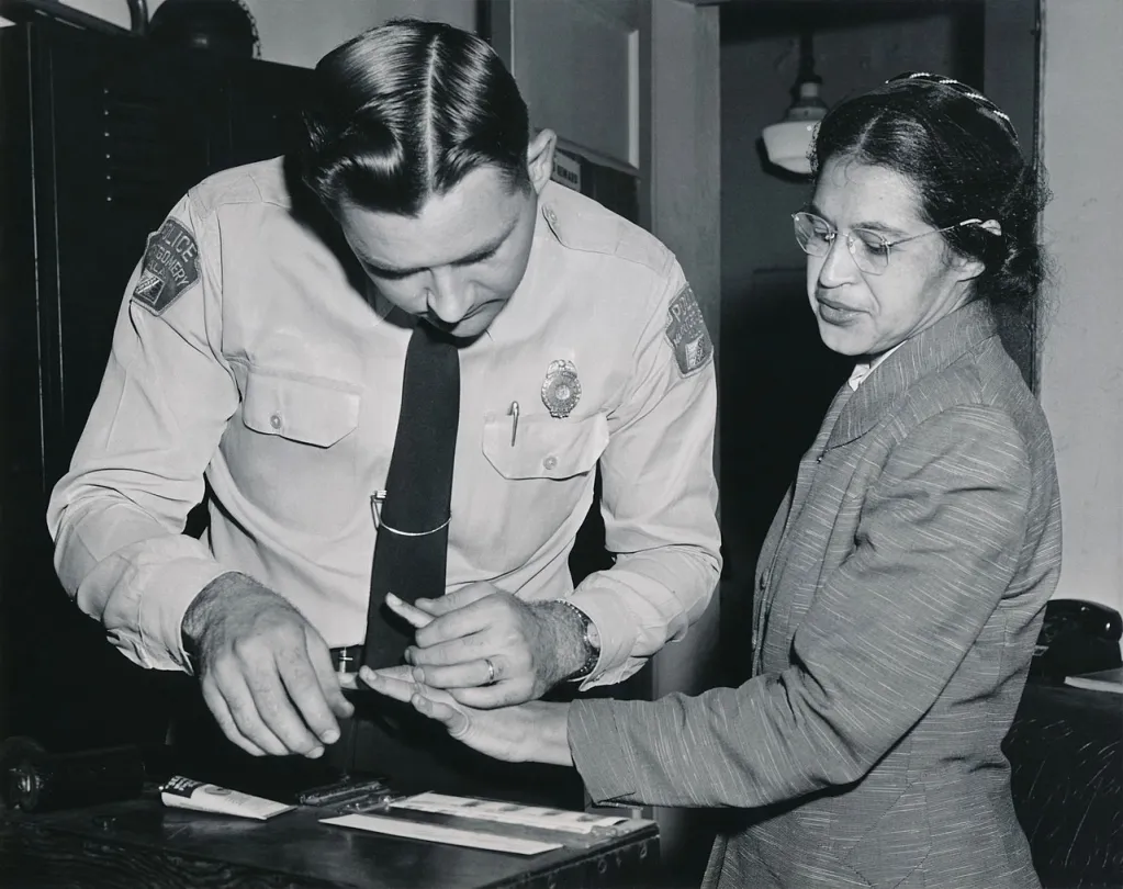 A historical photo of Rosa Parks, a Black woman wearing a suit, getting her fingerprints taken by a young white male cop.